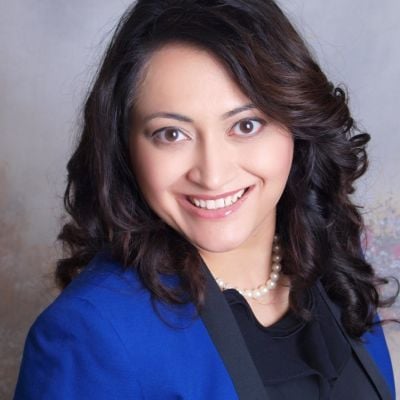 A smiling light-skinned female with dark brown hair wearing a cobalt jacket against a gray background