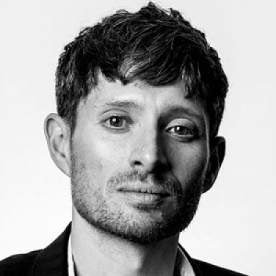 A black and white photo of a light-skinned male with a beard wearing a dark suit against a light background