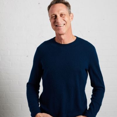 A smiling light-skinned male with gray hair wearing a navy sweater against a light gray background