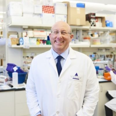 A smiling light-skinned male wearing glasses and a white lab coat over a button-up and tie against a pharmacy setting