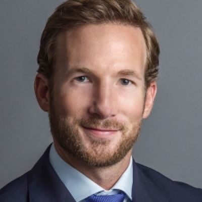 A smiling light-skinned male with light brown hair and a beard wearing a navy suit and tie against a gray background