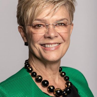 A smiling light-skinned female with short blonde hair wearing glasses and a green shirt against a light gray backdrop