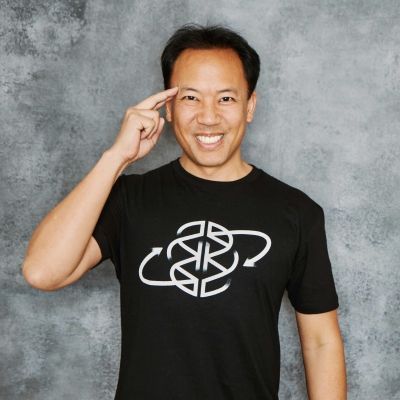 A smiling male of Asian heritage wearing a black tshirt with his hand by his temple against a gray marbled backdrop