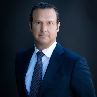 A light-skinned male with dark brown hair wearing a navy suit and tie against a dark gray background