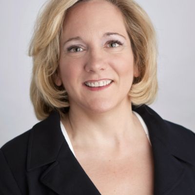 A smiling light-skinned female with short blonde hair wearing a black suit jacket against a light gray background