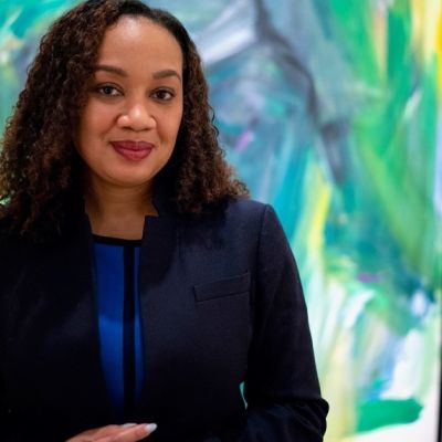 A smiling darker-skinned female with black hair wearing a cobalt shirt with a dark suit jacket against an abstract colorful background