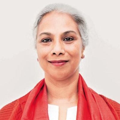 A smiling light-skinned female with gray hair wearing a coral shawl over a white shirt against a cream background