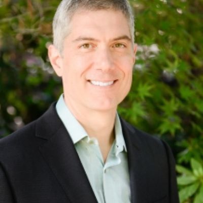 A smiling light-skinned male with gray hair wearing a black suit jacket and a light green button-up shirt against an outdoor backdrop