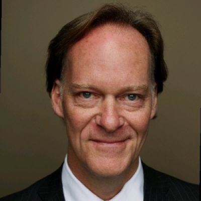 A light-skinned male-presenting person with dark brown hair wearing a black suit against a light brown background