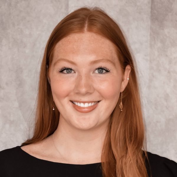 Smiling light-skinned, female-presenting individual with long red hair wearing a dark shirt on a brown backdrop.