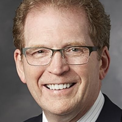 A smiling light-skinned male with red brown hair wearing glasses and a dark suit against a gray backdrop