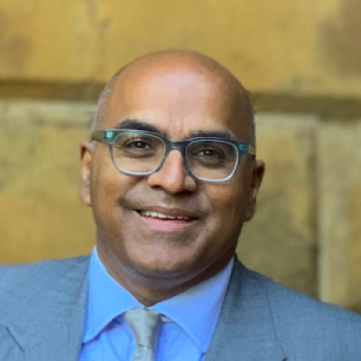 A smiling darker-skinned male wearing glasses and a light blue suit and tie against an ambiguous background