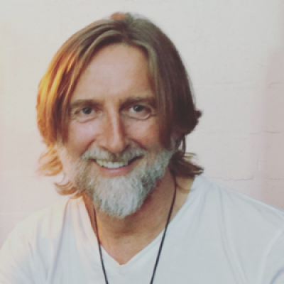 A smiling light-skinned male with gray hair and a beard wearing a white shirt against a light gray background
