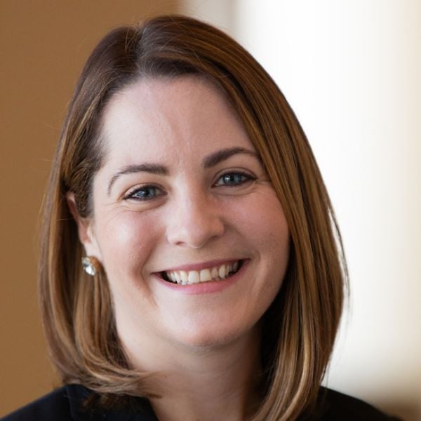 Smiling light-skinned, female-presenting individual with short hair wearing a dark shirt on a brown backdrop.