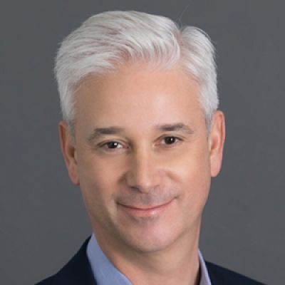 A smiling light-skinned male with white hair wearing a navy suit against a gray background
