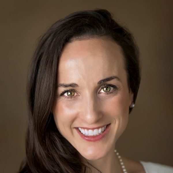 Smiling light-skinned, female-presenting individual with long dark hair wearing a white shirt on a brown backdrop.