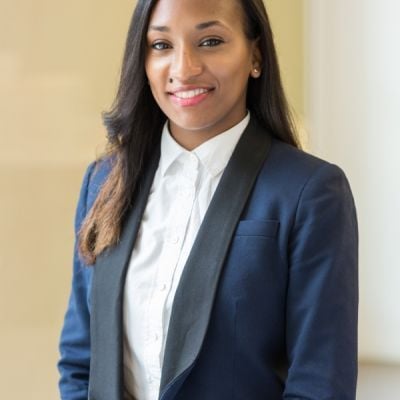 A smiling darker-skinned female with long black hair wearing a navy and black suit jacket over a white button-up shirt against a light brown background