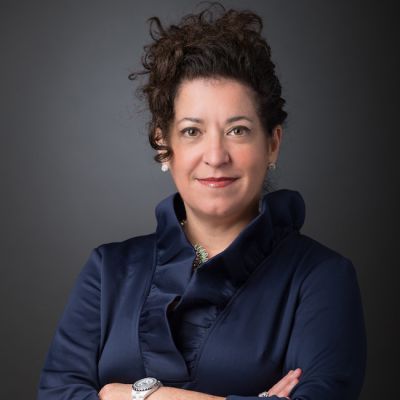 A light-skinned female with dark brown curly hair wearing a ruffled navy shirt against a dark gray background