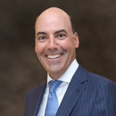 A smiling light-skinned male wearing a blue suit and tie against a dark gray background