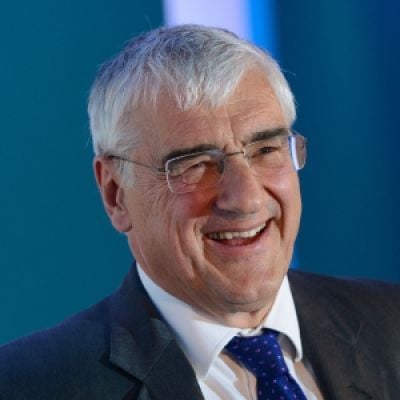 A smiling light-skinned male with white hair wearing glasses and a dark suit and tie against a blue background