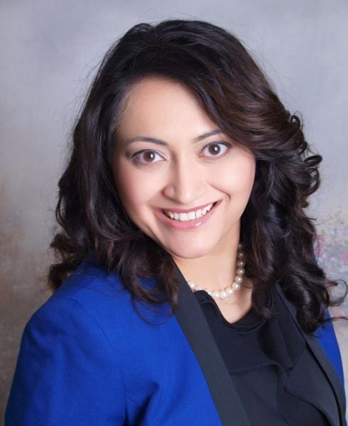 A smiling lighter-skinned woman with dark brown hair wearing a blue jacket over a black shirt against a gray background