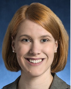 A smiling light skinned female with short red hair wearing a gray top against a blue background