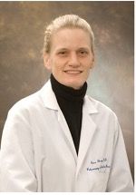 A smiling light-skinned female with blonde hair wearing a black turtleneck under a lab coat against a brown marbled background