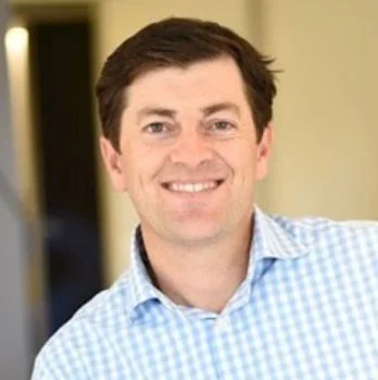 A smiling light-skinned male with dark brown hair wearing a checkered blue and white shirt against an ambiguous background