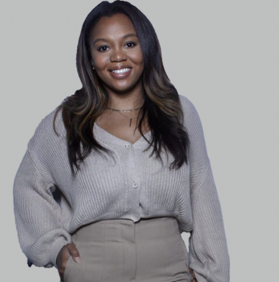 A smiling Black female with long black hair wearing a gray sweater over gray trousers stands with her hand in her pocket against a light gray background