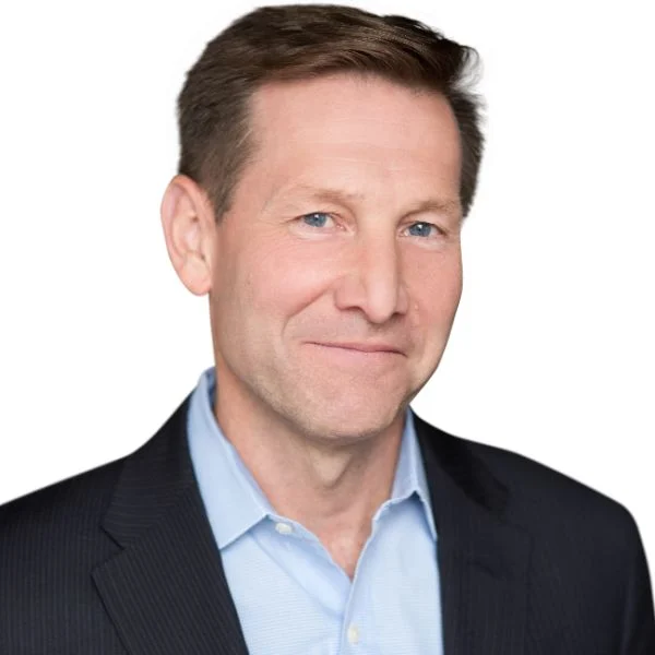 A smiling light-skinned male with brown hair wearing a dark suit jacket and a light blue button-up against a white background