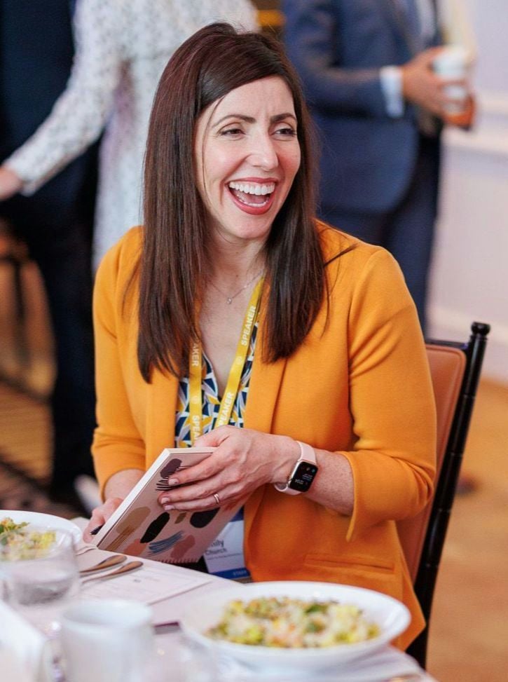 A smiling light-skinned female with brown hair wearing a yellow blazer over a patterned shirt against an ambiguous background