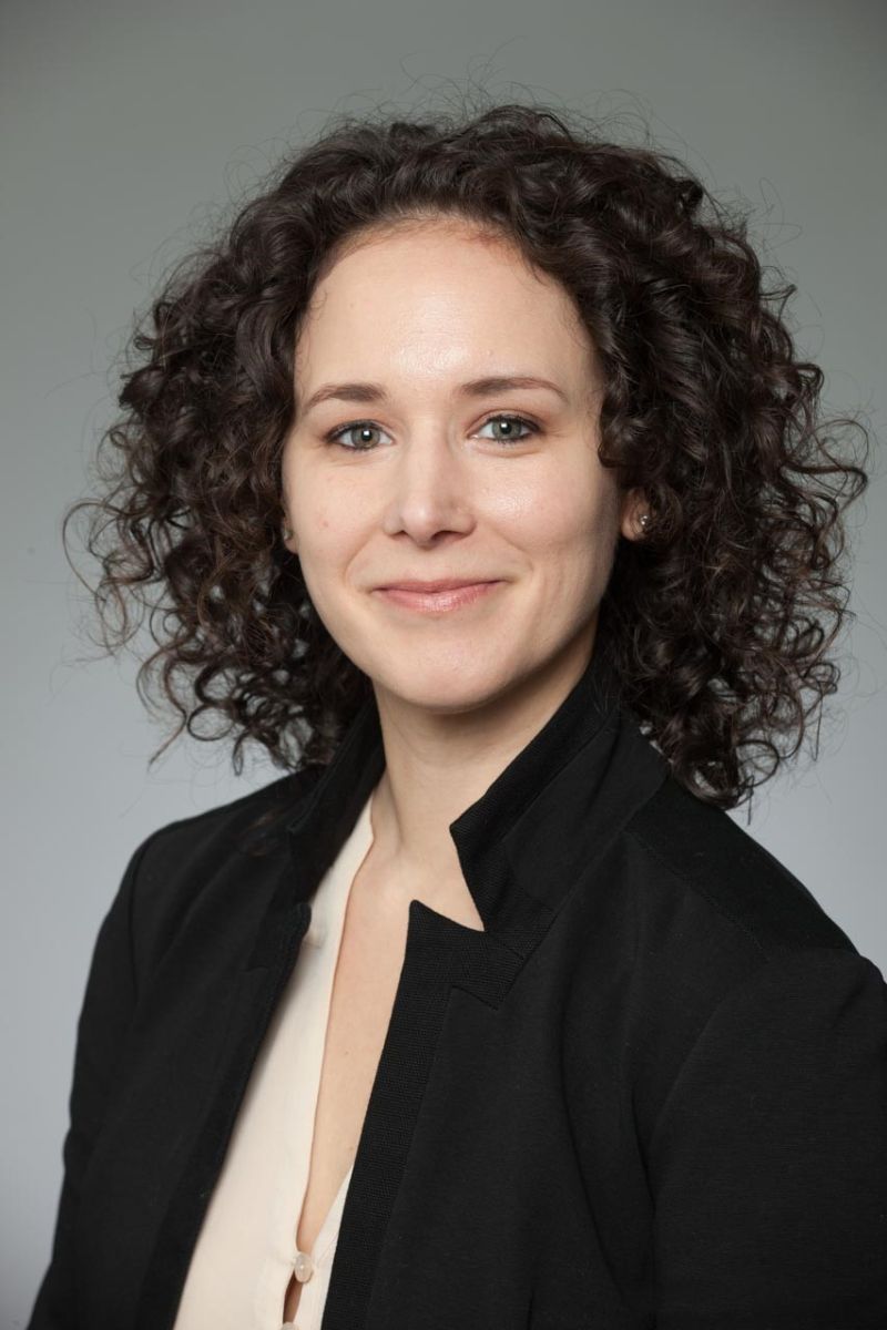 A woman with short, curly hair in a black blazer against a gray background.