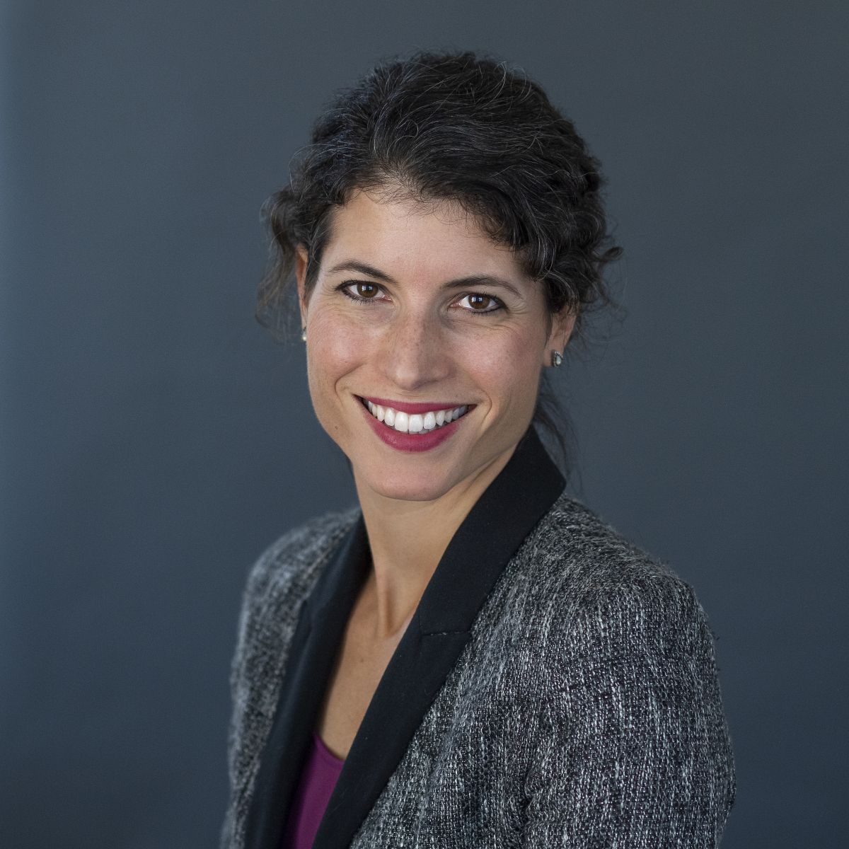 A smiling light-skinned female with dark grown hair wearing a gray jacket over a purple shirt against a gray background