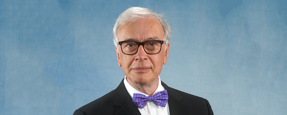 A light-skinned male with short white hair wearing a black suit and bowtie and glasses against a blue background