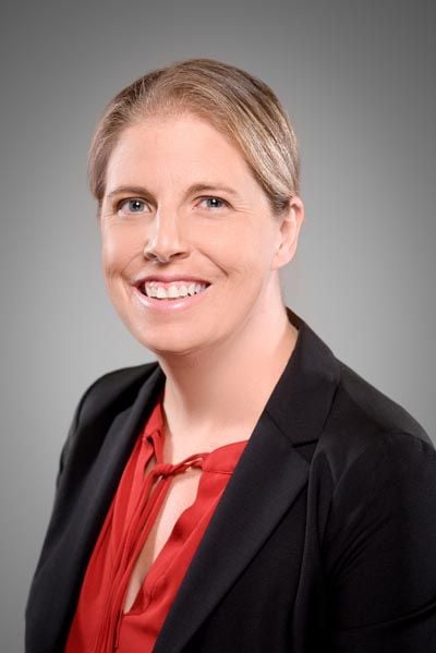 A smiling, light-haired woman wearing a red top and a black blazer against a gray background.