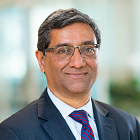 A smiling darker-skinned male with black and gray hair wearing a suit and tie against an ambiguous background