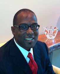 A smiling headshot of an adult man with brown skin, buzzed hair, and rectangular glasses. He is wearing a suit with a red tie.