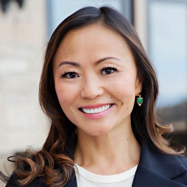 A headshot of a smiling adult woman who is Asian in appearance. She is wearing jade earrings and her hair is brown and shoulder-length. She is also wearing a white undershirt and a blue collared jacket.