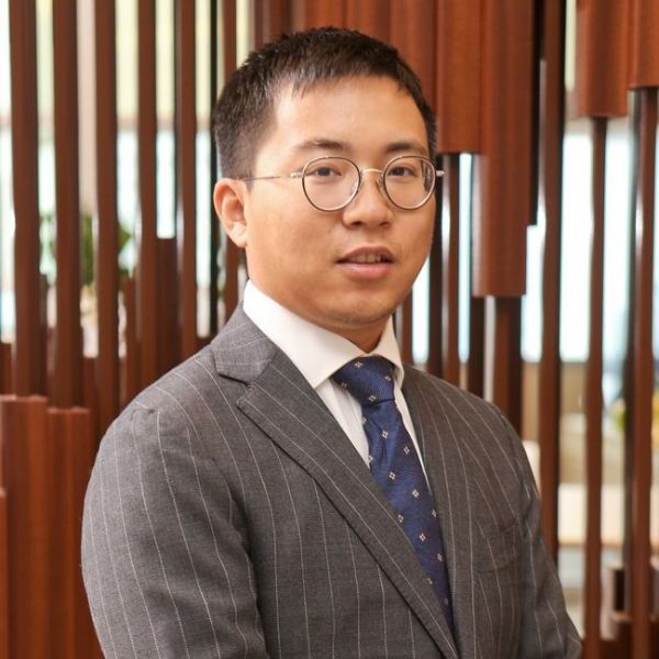 An image of an adult man who is Asian in appearance, with short black hair, wearing round clear glasses, and wearing a gray suit with a blue tie.