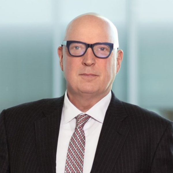 An image of a bald man with white skin, wearing thick black framed glasses and a suit and tie. He is looking seriously at the camera. 