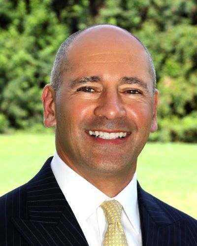 An image of an adult man with white tanned skin and a bald head, who is smiling at the camera. He is wearing a suit with a yellow tie.