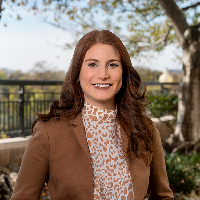 A smiling light-skinned female-presenting person named Chelsea Wendel with red hair wearing a brown cheetah print shirt against a blurred background.