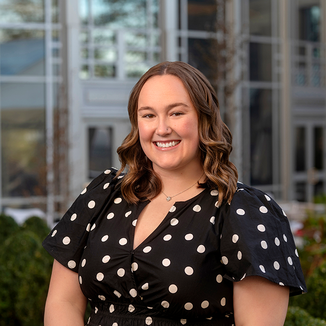 An image of a woman with white skin named Gretchen Jacobs. She has shoulder-length brown hair and is wearing a dark shirt with white polka dots. She is smiling at the camera with a building in the background.