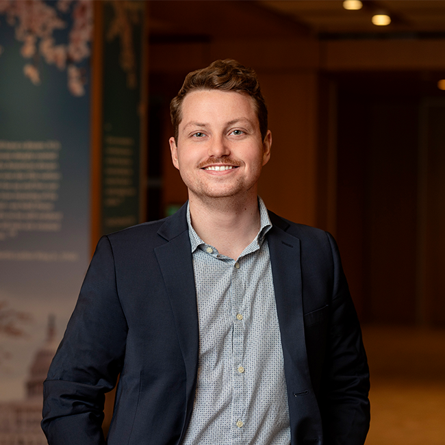 A smiling light-skinned man named Zachary Walsh with short brown hair, wearing a navy blue blazer over a grey button-down shirt, and standing in a room with dark wood details.