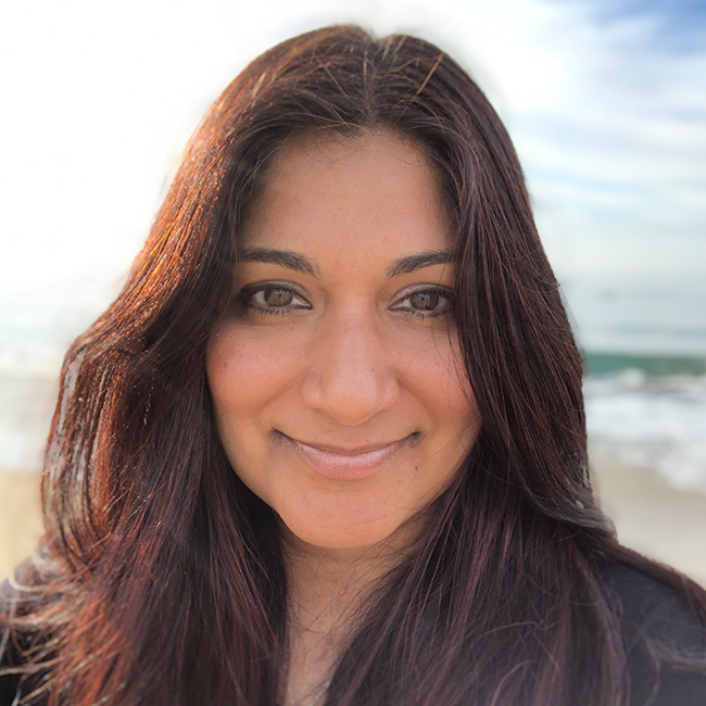 An image of a woman named Ani Chahal Honan. She has tan skin, brown eyes, and long brown hair. She is smiling at the camera and wearing a black shirt against an ambiguous outdoor background.
