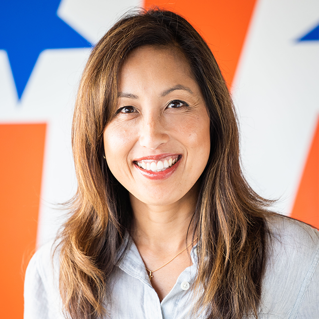 A headshot of a woman who is Asian in appearance with white skin, brown eyes, shoulder-length brown hair, and a flag background with orange and white stripes and blue stars. She is wearing a gray buttoned up shirt and smiling at the camera.