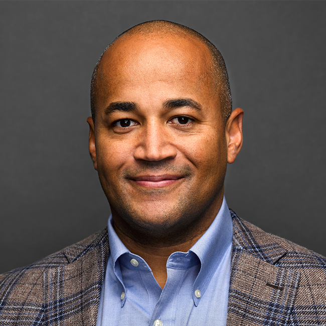An image of a man named Dante Disparte. He has light brown skin, a bald head, brown eyes, and he is wearing a plaid blazer with a blue shirt and smiling at the camera against a gray background.