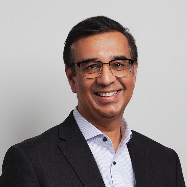 An image of a man named Kamal Bhatia. He has is standing against a gray background, wearing a suit and collared shirt and glasses, and smiling at the camera. He has short dark brown hair.