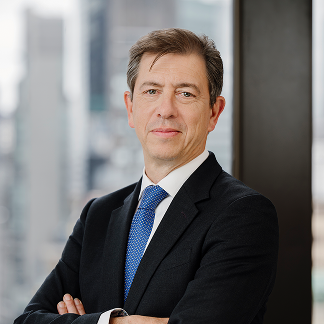 An image of a man named Mathias Burghardt. He is standing against a city background with his arms crossed and looking at the camera. He is wearing a dark suit with a blue tie and has white skin and short brown hair.