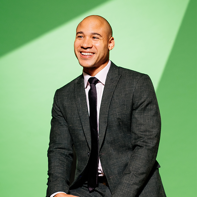 An image of a man named Michael Ellison. He is Asian in appearance and has tan white skin and a bald head. He is smiling and staring into the distance against a light green background and wearing a dark suit and tie. 